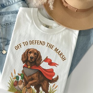 May include: A white t-shirt featuring a brown dog wearing a red cape and chainmail armour, holding a small red flag. The text reads OFF TO DEFEND THE MARSH above the dog, with two ducks and bulrushes at the bottom. Light blue jeans and a tan hat.