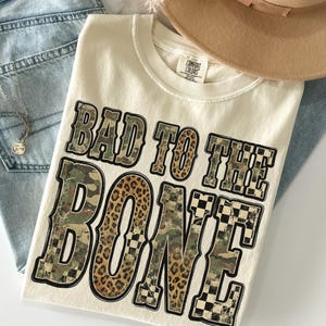 May include: Cream-colored t-shirt with the phrase "BAD TO THE BONE" in a bold, multi-textured design. The text incorporates camouflage, leopard print, and checkered patterns. The shirt is laid on top of blue jeans and a tan hat.