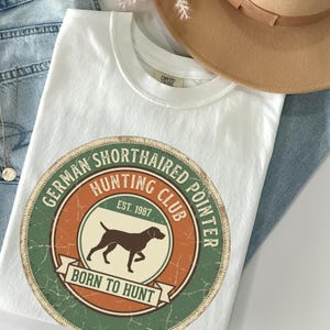 May include: White t-shirt with a circular graphic featuring a German Shorthaired Pointer dog, the text "German Shorthaired Hunting Club EST. 1987" and "Born to Hunt". The shirt is laid on top of blue jeans and a tan hat.