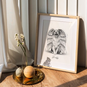 May include: A black and white drawing of a dog with wings, framed in a light wood frame. The dog is smiling and the drawing is titled "Buddy".
