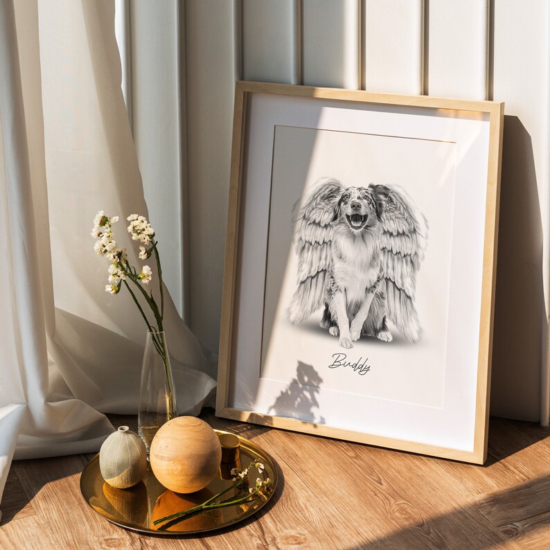 May include: A black and white drawing of a dog with wings, framed in a light wood frame. The dog is smiling and the drawing is titled "Buddy".