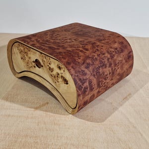 Könnte beinhalten: Eine handgefertigte Holzkiste mit geschwungenem Design. Die Kiste hat eine Schublade aus hellem Holz und eine Oberseite mit einem rötlich-braunen Maserungsmuster. Die Kiste steht auf einer hellen Holzoberfläche.