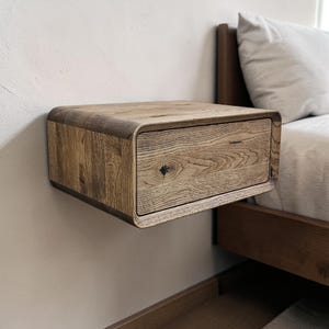 May include: A floating wooden nightstand with a single drawer. The nightstand is made of light-colored wood with visible grain and a rounded top and front edges. It is mounted on a white wall near a bed.