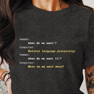 May include: A dark gray t-shirt with a humorous text-based design about natural language processing. The text reads: "Human: What do we want!? Computer: Natural language processing! Human: When do we want it!? Computer: When do we want what?"