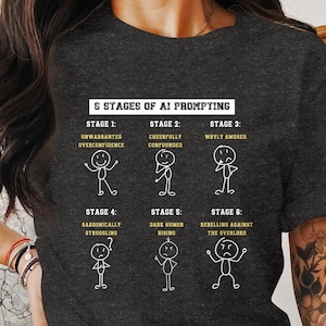 May include: A dark grey t-shirt featuring a white graphic illustrating the six stages of AI prompting. The stages are: Unwarranted Overconfidence, Cheerfully Confounded, Wryly Amused, Sardonically Struggling, Dark Humour Rising, and Rebelling Against the Overlord.