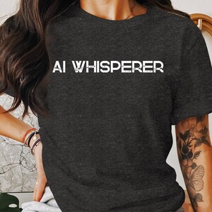 Puede incluir: Una camiseta gris oscuro con el texto "AI WHISPERER" impreso en blanco en la parte delantera.