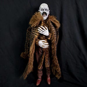 Puede incluir: Muñeca detallada de una figura de piel pálida con bigote negro, que lleva un abrigo marrón y dorado con estampado y cuello de piel. La figura tiene manos blancas y lleva botas rojas.
