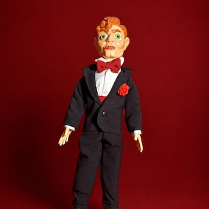 May include: A ventriloquist dummy with orange hair and green eyes, wearing a black suit jacket, black trousers, a white shirt, a red bow tie, and a red flower lapel pin. The figure has a pale complexion and a moulded facial expression.