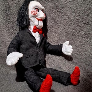 Puede incluir: Muñeco de un personaje de la serie de películas de terror Saw. El muñeco tiene una cara blanca con mejillas en espiral rojas, labios rojos y cabello negro. Viste un traje negro, camisa blanca, pajarita roja, guantes blancos y zapatos rojos.