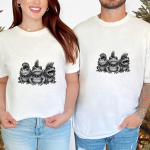 May include: White t-shirts featuring a black and white illustration of three punk rock frogs. The frogs have spiky mohawk hairstyles and are wearing studded collars. The shirts are a casual, unisex style.