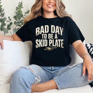 Könnte beinhalten: Schwarzes T-Shirt mit dem Text "BAD DAY TO BE A SKID PLATE" in verblichener weißer Schrift. Das Shirt zeigt eine Grafik einer Unterfahrschutzplatte. Das Model trägt hellblaue Jeans.