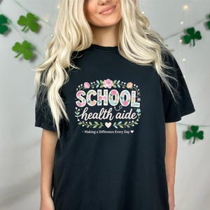 Puede incluir: Camiseta negra con la frase "SCHOOL health aide" en un diseño floral. El texto "Making a Difference Every Day" está impreso debajo. La camiseta tiene cuello redondo y mangas cortas.