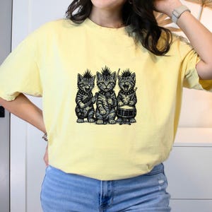 May include: A light yellow t-shirt featuring a black and white illustration of three punk rock cats. One cat plays a guitar, another sings into a microphone, and the third plays a drum set. The cats have mohawk haircuts and studded collars.