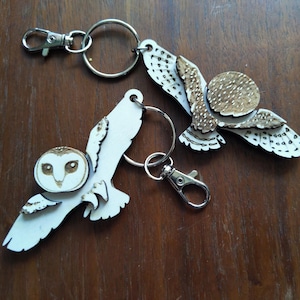 May include: Two wooden owl keychains with silver-colored clasps and key rings. The owls are white with brown and tan accents, and are in a flying pose. The keychains are on a dark brown wooden surface.
