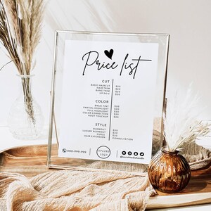 Op de afbeelding: Een witte prijslijst voor een kapsalon met een hartje en de tekst "Price list" bovenaan. De lijst bevat prijzen voor kapsels, haar knippen, kleurdiensten en stylingdiensten. De lijst is ingelijst in een zilveren lijst en staat op een houten tafel met een vaas met gedroogde bloemen en een kleine vaas met een bruine bloem.