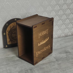 Könnte beinhalten: Eine Holzkiste mit einem Scharnierdeckel, auf dem eine Silhouette von Figuren aus "Der Hobbit" und "Der Herr der Ringe" zu sehen ist. Die Kiste ist aus dunkelbraunem Holz gefertigt und hat eine natürliche Holzoptik.