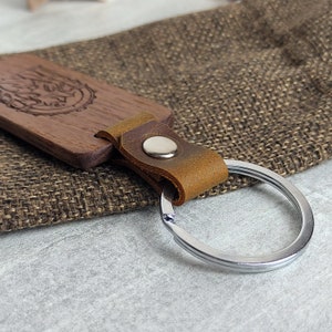 May include: A wooden keychain with a leather strap and a silver ring. The wood is dark brown and has a carved design. The leather strap is brown and has a metal button.