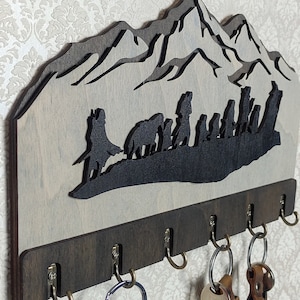 Op de afbeelding: Houten sleutelhanger met een silhouet van een bergketen en figuren die er voor lopen. De sleutelhanger heeft zes haken om sleutels aan te hangen.
