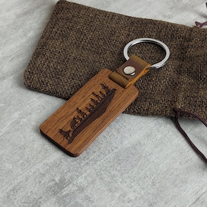 May include: A wooden keyring with a brown leather strap and a silver key ring. The keyring features a laser-engraved design of a group of trees on a hill.