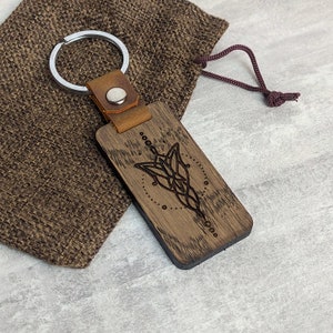 May include: A rectangular wooden keyring with a silver key ring and a brown leather strap. The wooden part has a detailed engraved design. The keyring is next to a brown burlap bag.