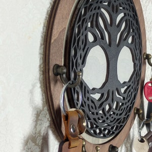 Op de afbeelding: Een ovale houten sleutelhouder met een lasergesneden levensboomontwerp. De sleutelhouder heeft een donkerbruin frame en metalen haken voor sleutels. Er hangen verschillende sleutels aan de haken.