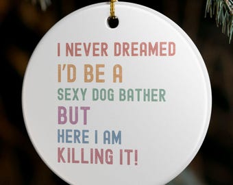 May include: A white, round Christmas ornament with the text "I NEVER DREAMED I'D BE A SEXY DOG BATHER BUT HERE I AM KILLING IT!" in various colors. The ornament hangs from a gold string against a blurred background of Christmas tree branches and lights.
