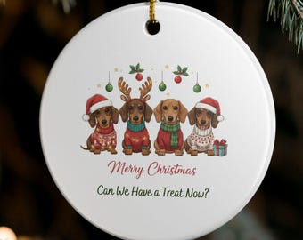 以下が含まれることがあります： 白い丸いクリスマスオーナメント。4匹のダックスフンドが、お祝いのセーターと帽子を着用しています。「Merry Christmas」と「Can We Have a Treat Now?」の文字が赤と緑で書かれています。金色の紐で吊り下げられており、背景には松の枝があります。