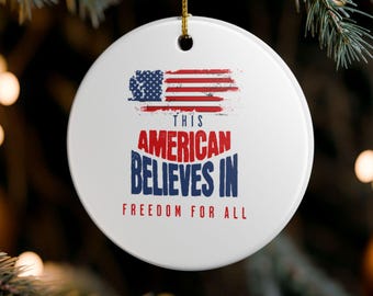 May include: A white, round Christmas ornament with a patriotic design. It features a stylized American flag and the text "THIS AMERICAN BELIEVES IN FREEDOM FOR ALL" in red and blue. The ornament hangs from a gold string.