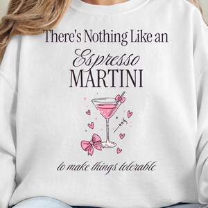 Könnte beinhalten: Weißes Sweatshirt mit dem Text "There's Nothing Like an Espresso Martini" über einer rosa Cocktail-Illustration mit Herzen und einer Schleife. Der Satz "to make things tolerable" steht darunter. Das Sweatshirt wird von einer Person getragen.