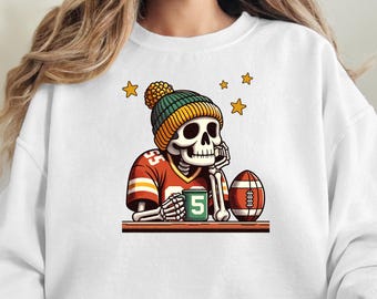 May include: White sweatshirt featuring a cartoon skeleton wearing a football jersey and a beanie. The skeleton is holding a mug with the number 5 and resting its head on its hand. A football and stars are also present.