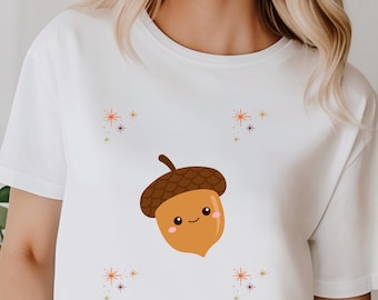 Puede incluir: Camiseta blanca con un diseño de bellota de dibujos animados sonriente. La bellota es marrón y beige, con un pequeño tallo. La camiseta también tiene pequeños diseños de estrellas en las esquinas.