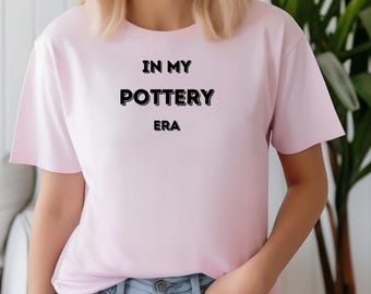 May include: A pale pink t-shirt featuring the black text "IN MY POTTERY ERA". The short-sleeved top is worn with blue jeans. The image shows a person smiling.
