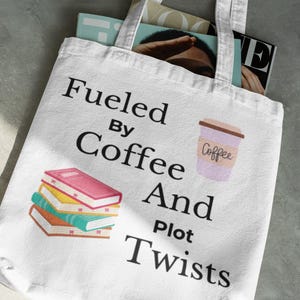 May include: White tote bag with the text "Fueled By Coffee And Plot Twists." The bag features illustrations of a stack of books and a coffee cup. Magazines are visible inside the bag.