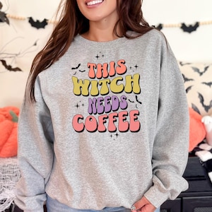 Op de afbeelding: Een grijs sweatshirt met een kleurrijke afbeelding waarop staat "This Witch Needs Coffee". De afbeelding is in retro-stijl met een roze, gele en paarse kleurstelling.