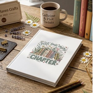 May include: A white hardcover journal featuring a colorful illustration of stacked books, flowers, and the text "Just one more CHAPTER" on the front cover. The journal is placed on a wooden surface alongside a mug, books, and floral accents.