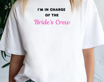 Puede incluir: Camiseta blanca con texto negro que dice "I'm in charge of the Bride's Crew". La palabra "Bride's" está en rosa.