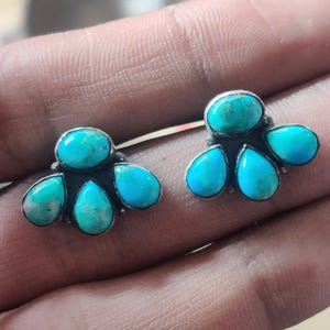 May include: A pair of turquoise stud earrings. Each earring features a central oval turquoise stone surrounded by four teardrop-shaped turquoise stones, set in a silver-toned metal. The earrings have a floral or petal-like design.