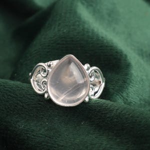 Könnte beinhalten: Ein silberner Ring mit einem tropfenförmigen Rosenquarz-Edelstein, der in einem filigranen Design gefasst ist. Der Ring wird auf einem grünen Samt-Hintergrund präsentiert.