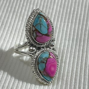Könnte beinhalten: Ein silberner Ring mit zwei tropfenförmigen Steinen. Jeder Stein hat eine türkisfarbene und magenta Farbgebung mit strukturiertem Aussehen. Der Ring hat einen detaillierten, perlenbesetzten Rand.