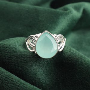 Könnte beinhalten: Ein silberner Ring mit einem hellblauen, tropfenförmigen Edelstein, der in einem filigranen Design gefasst ist.