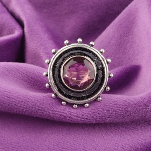 Könnte beinhalten: Ein silberner Ring mit einem großen, runden, facettierten, lila Edelstein in der Mitte. Der Edelstein ist von einem schwarzen Band und einem silbernen Band mit kleinen, runden silbernen Akzenten umgeben. Der Ring ist vor einem violetten Samthintergrund platziert.