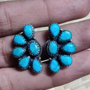 May include: A pair of silver-toned stud earrings, each featuring a cluster of turquoise teardrop-shaped stones. The earrings are set in a floral design, with each stone surrounded by a silver border. The turquoise stones have a vibrant, light blue color.