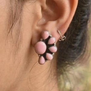 Könnte beinhalten: Nahaufnahme eines Ohrs mit einem Cluster-Ohrstecker. Der Ohrring hat ein florales Design mit mehreren hellrosa, ovalen Steinen, die in einen silbernen Metallrahmen gefasst sind. Ein kleiner silberner Ohrstecker ist ebenfalls sichtbar.