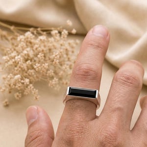 Könnte beinhalten: Ein silberner Ring mit einem rechteckigen, horizontal ausgerichteten schwarzen Stein, der in ein glattes Metallband gefasst ist. Der Ring hat eine flache, polierte Oberfläche und ein klares, geometrisches Design, das auf dem Finger aufliegt.