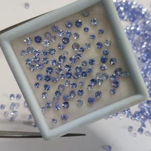 May include: A collection of small, round, light blue gemstones. Many are contained within a white rectangular tray, while others are scattered around. The gemstones have multiple facets, reflecting light. A pair of tweezers is visible.