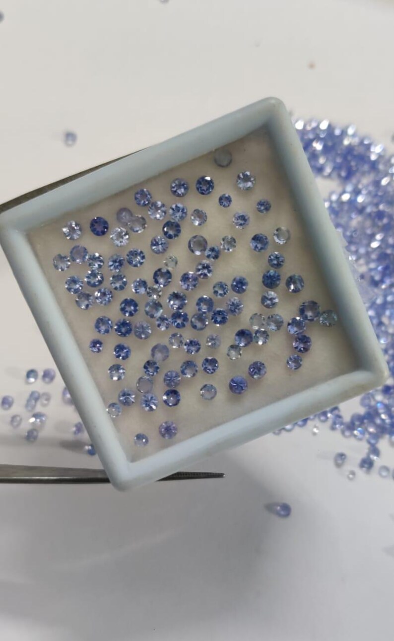 May include: A collection of small, round, light blue gemstones. Many are contained within a white rectangular tray, while others are scattered around. The gemstones have multiple facets, reflecting light. A pair of tweezers is visible.