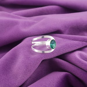 May include: A silver ring with a large, oval-shaped, emerald-green gemstone. The ring has a split band design, with two parallel bands curving around the finger. The ring is set against a purple fabric background.