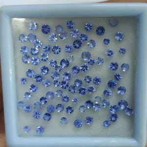 May include: A collection of small, round, blue gemstones of varying sizes are displayed inside a light blue square container. The gemstones have a faceted cut, reflecting light. The container is filled with many gemstones.