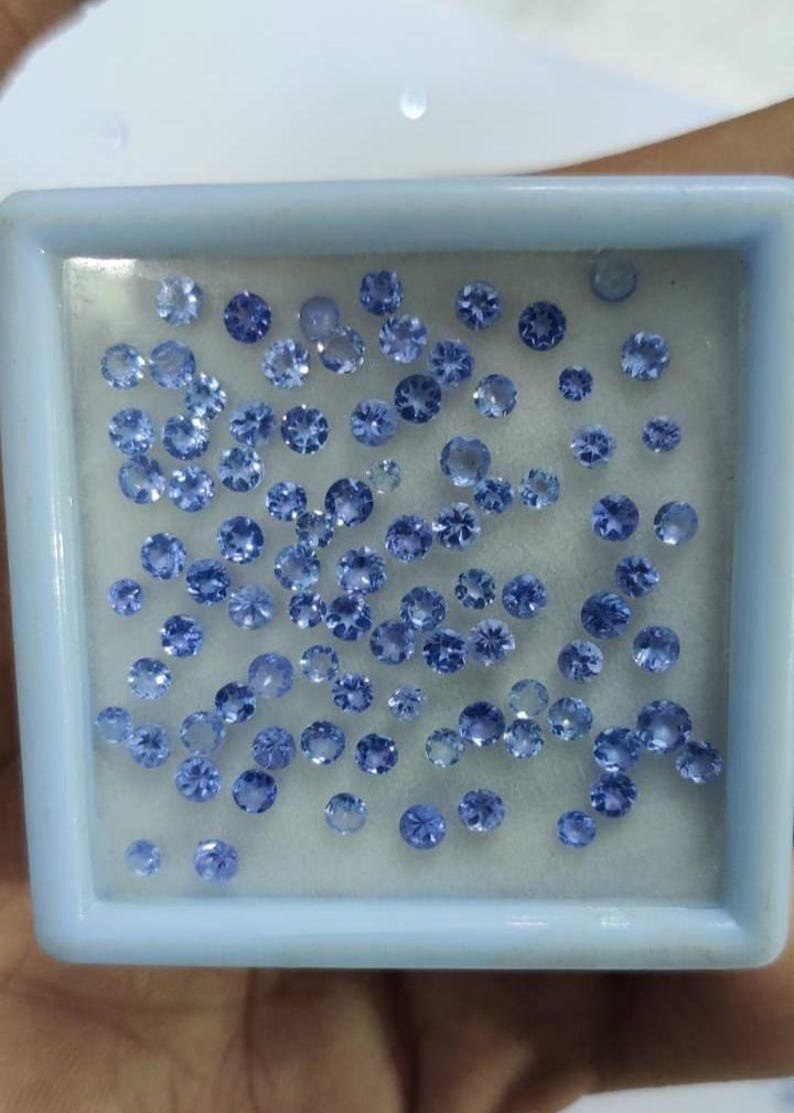 May include: A collection of small, round, blue gemstones of varying sizes are displayed inside a light blue square container. The gemstones have a faceted cut, reflecting light. The container is filled with many gemstones.
