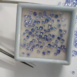 May include: A collection of small, round, light blue gemstones displayed in a white square container. The gemstones are of varying sizes and are scattered around the container. Tweezers are visible in the image, suggesting the gemstones are being handled.